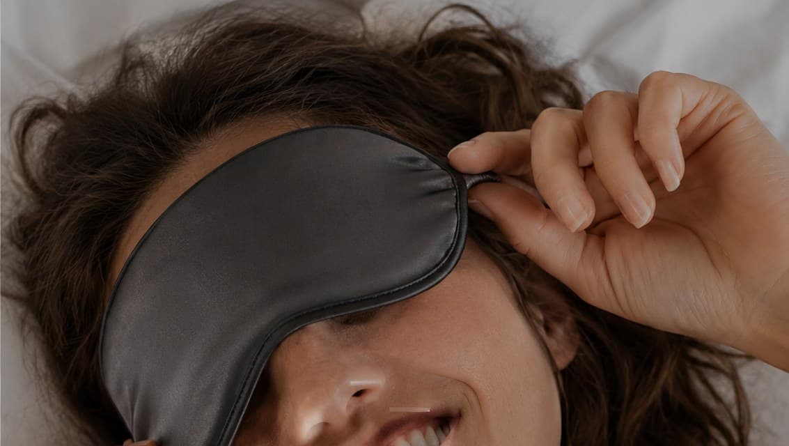 A woman lifting a silk sleep mask, resting on a pillow
